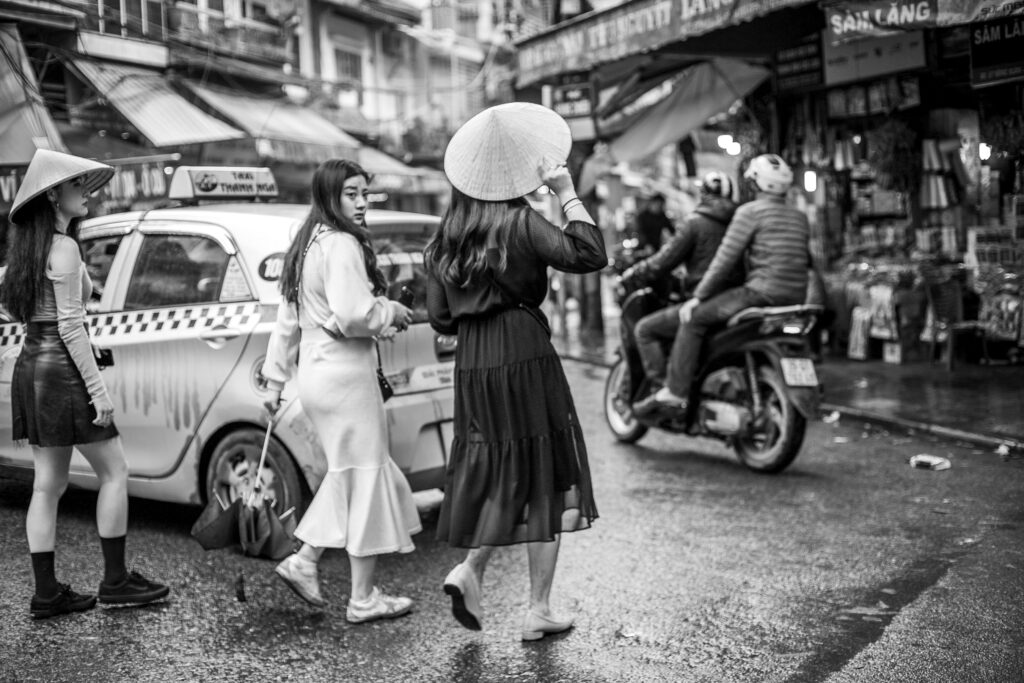 Black and white photo of three Vietnamese girls crossing the street. Girl in white dress looks back. Vietnam street photography